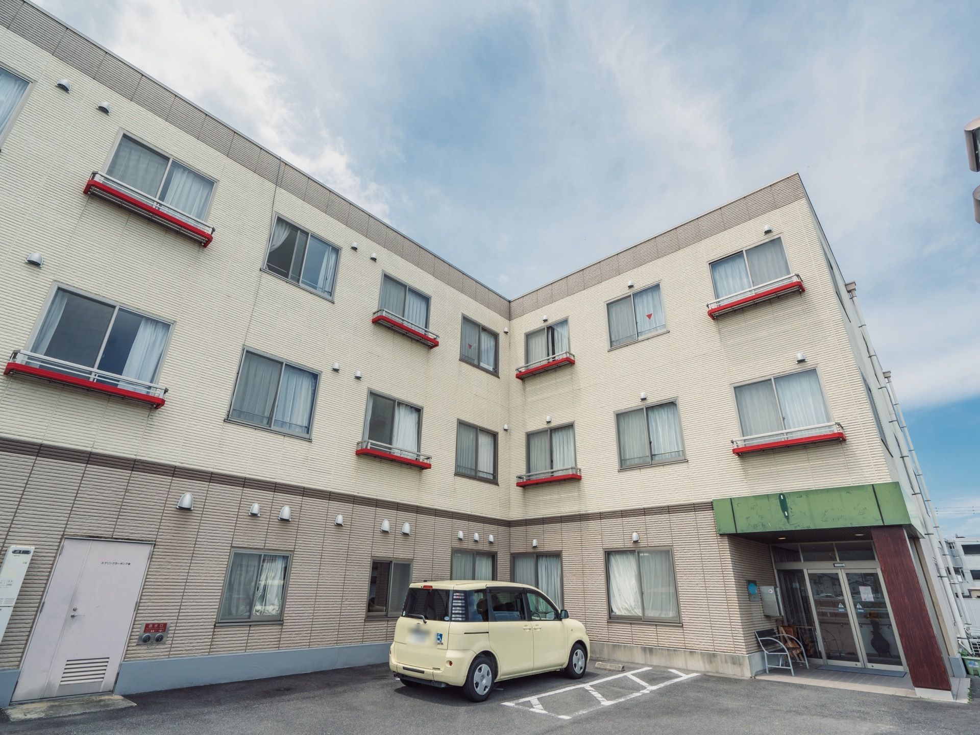 施設は３階建てで、窓がたくさん付いている。建物の前には白い車が停まっており、玄関の側にはベンチが置かれている。