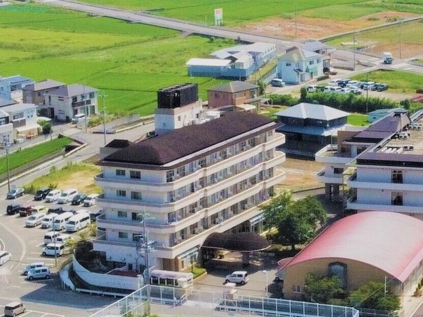 ブラウンの屋根が付いた5階建ての大きな建物のそばには広い駐車場があり、ピンクの楕円形の屋根が付いた体育館のような建物もある。