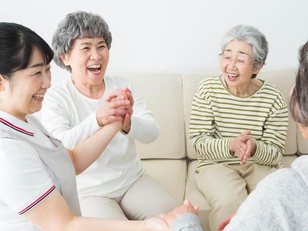 高齢者2名とスタッフが手を握りながら談笑している様子。施設内の会話の一画面