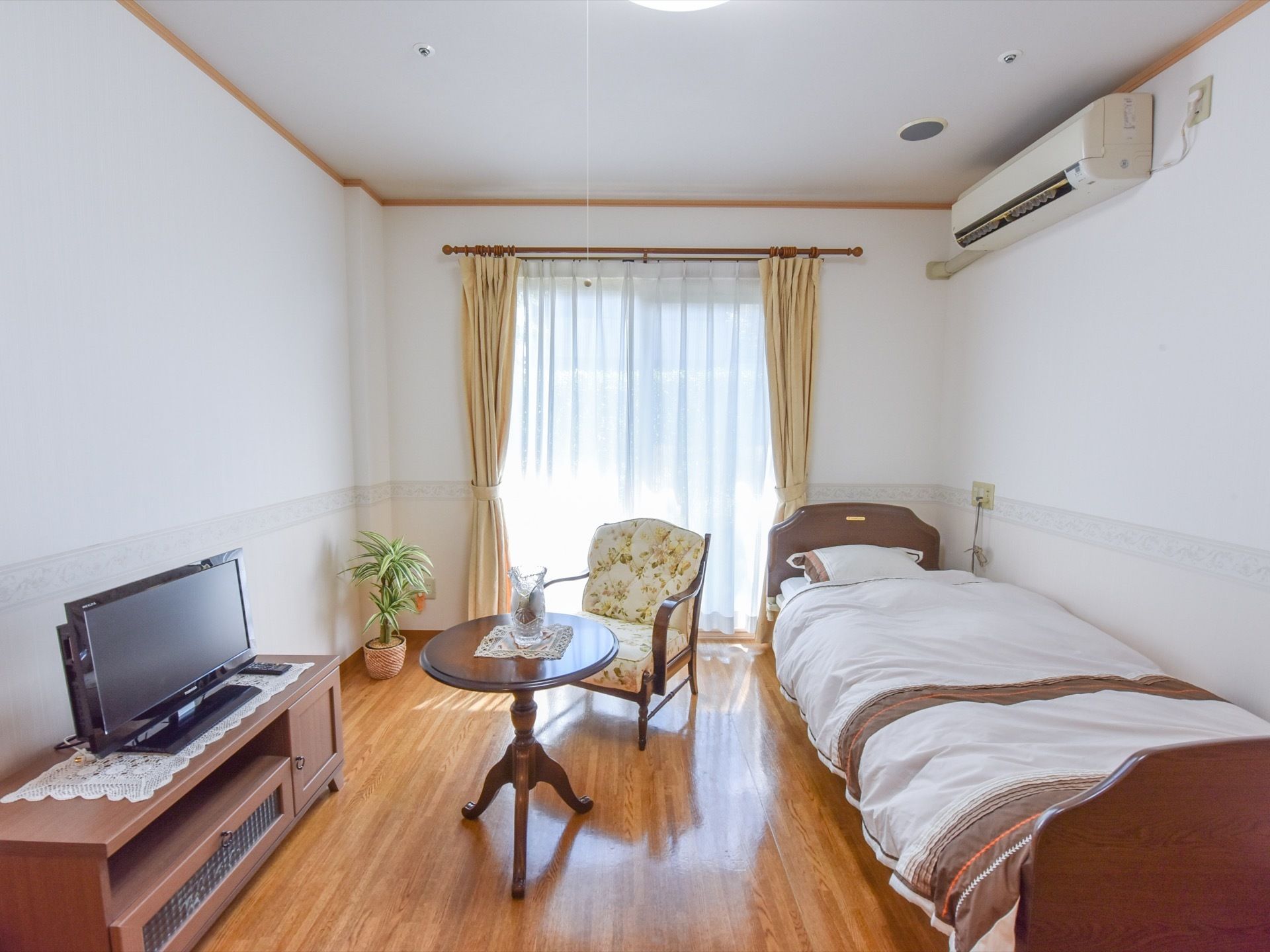 入居者様用の居室内には一通りの家具が揃っている。中央にはイスとテーブルがあり、ベッドやテレビ、観葉植物や空調機も設置されている。