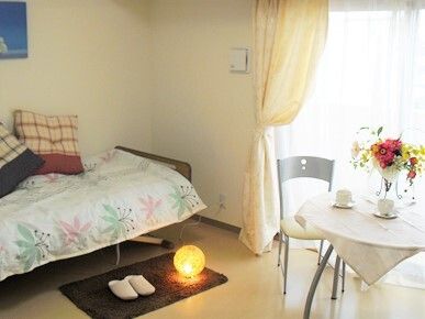 花柄の寝具がセットされたベッドの上にはクッションが二つ置かれ、マットの上にはスリッパがあり、テーブルには赤や黄色の花が生けられている。