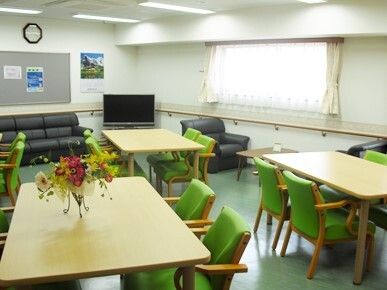 グリーンの椅子がセットされた木製のダイニングテーブルが複数並んだ食堂には、テレビや黒いソファも置かれ、壁にはカレンダーもある。