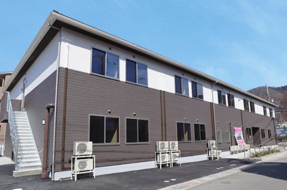 2階建ての建物は横に長く、1階部分と2階部分では外壁の色が異なる。側面には屋外階段が付いていて、周囲には山も見える。