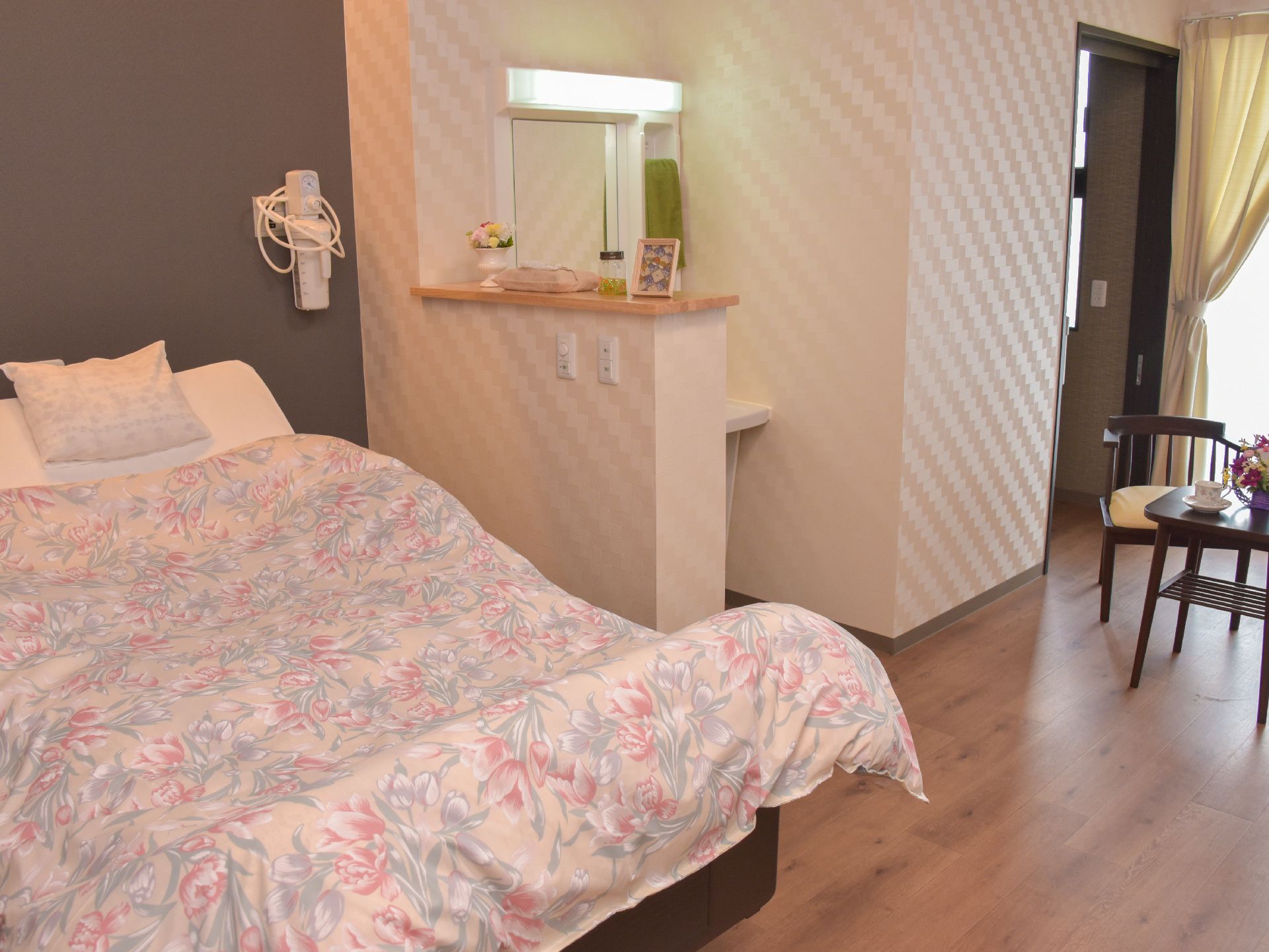 柄のついた壁紙の部屋で、電動ベッドがおいてあるほか、仕切りがありその横に洗面台が設置してある。大面鏡が見える。