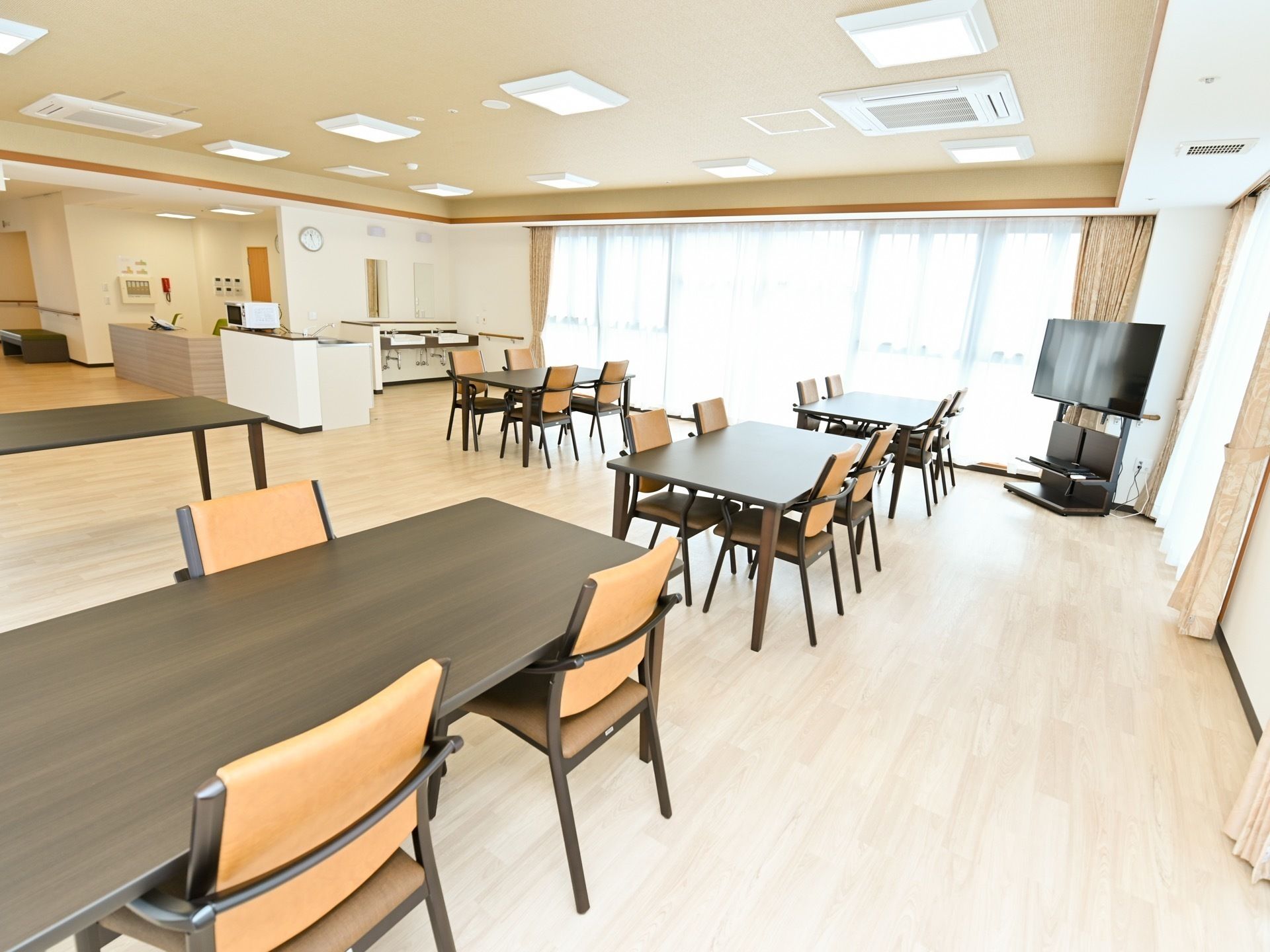 大きな窓から陽が射し込む広い食堂に、ダイニングテーブルと椅子が並んでいる。コーナーには、テレビが置かれている。