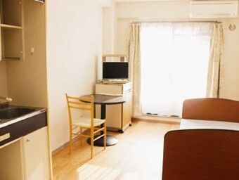 レースカーテンとカーテンが付いた大きな窓があるフローリングの部屋。二人用のテーブルセットやベッドがあり、ミニキッチンも付いている。