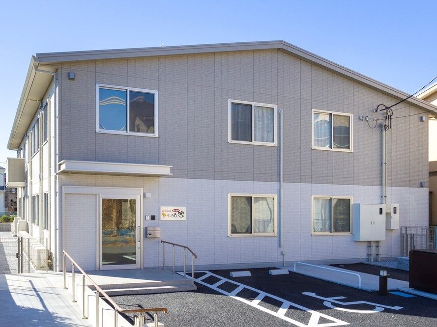 2階建ての建物。駐車スペースがあり、玄関には手すり付きのスロープが付き、車いすの高齢者と付き添いの人がいる。