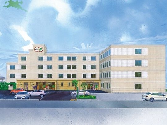 広い駐車場には複数の車が止まっている。ベージュの外壁の4階建ての建物の一番上には住宅のマークがある。