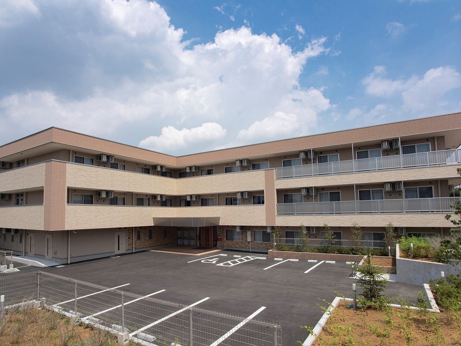 ３階建ての建物はエル字型で、各フロアにベランダがあり、ベージュとブラウンでまとめられ、広い駐車場がある。