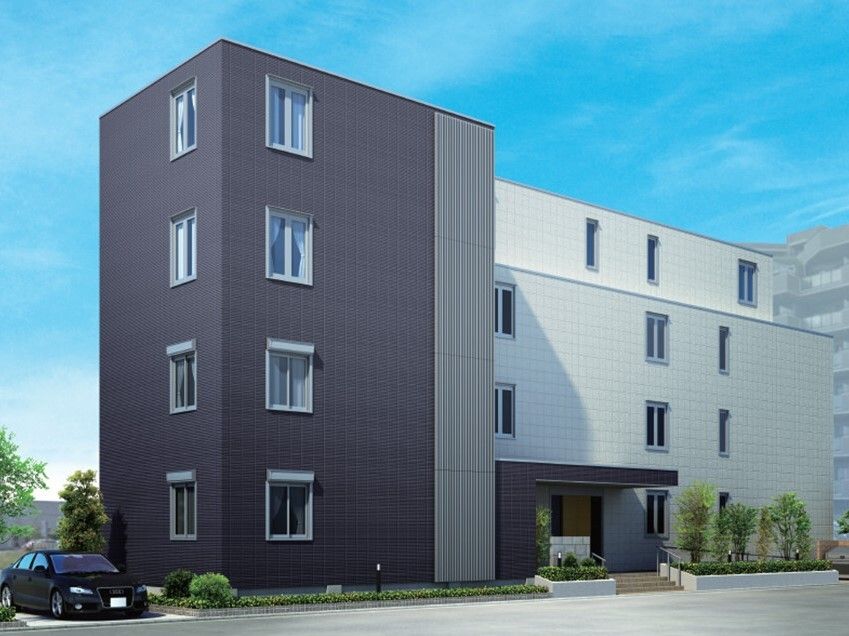 ４階建ての建物の玄関には手すり付きの階段とスロープがあり、屋根も付いている。白とグレーの外壁。