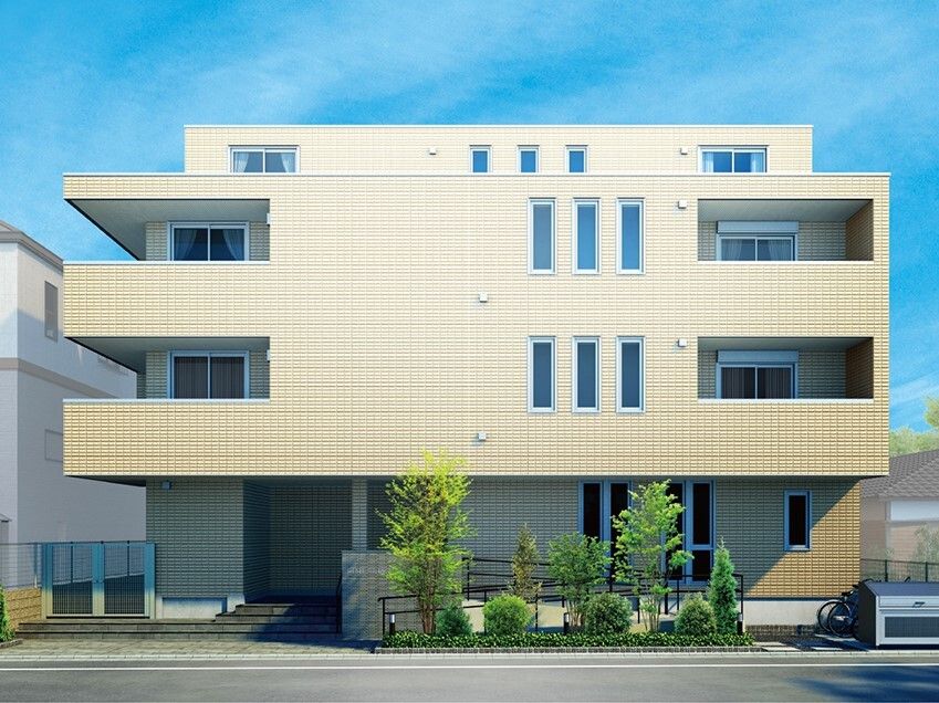 ベージュ系の４階建ての建物の周りには草木が植えられ、１階部分は少し面積が狭く、玄関の周りには手すり付きのスロープと階段がある。
