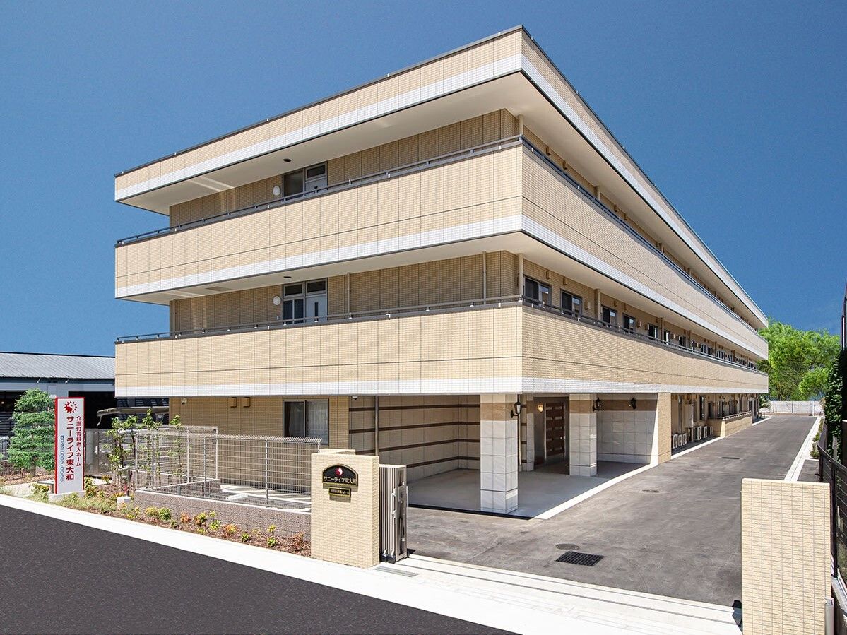 ベージュ系の３階建ての建物は横に長く、各フロアにベランダがあり、門のそばにはフェンスもある。