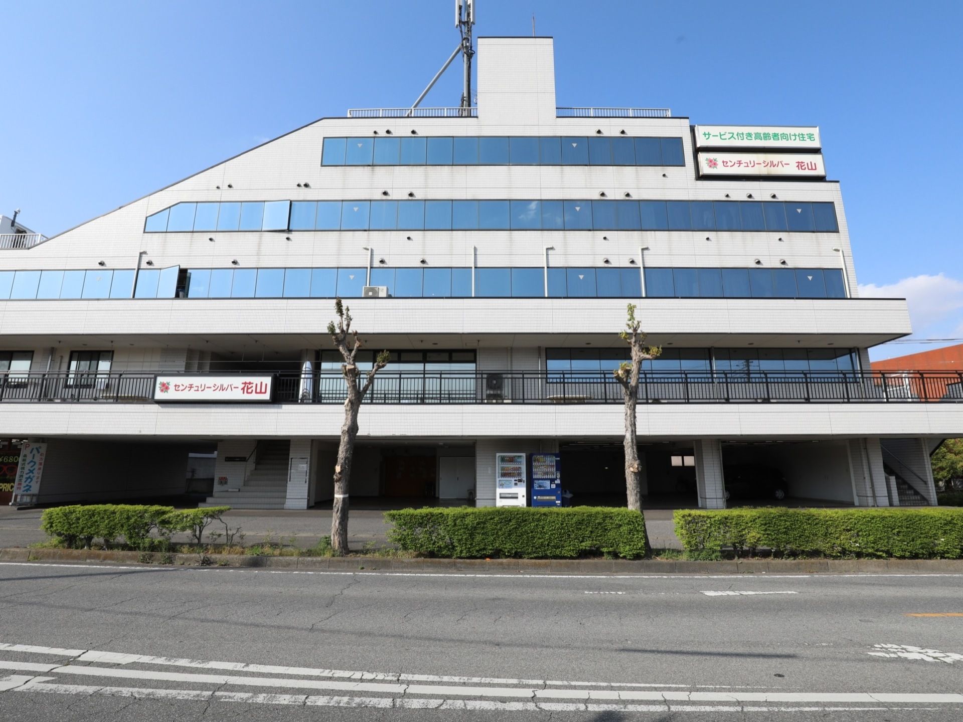 施設の外観。道路沿いに、植物や木が植えられている。1階には階段が2箇所あり、中央に自動販売機が2機設置されている。