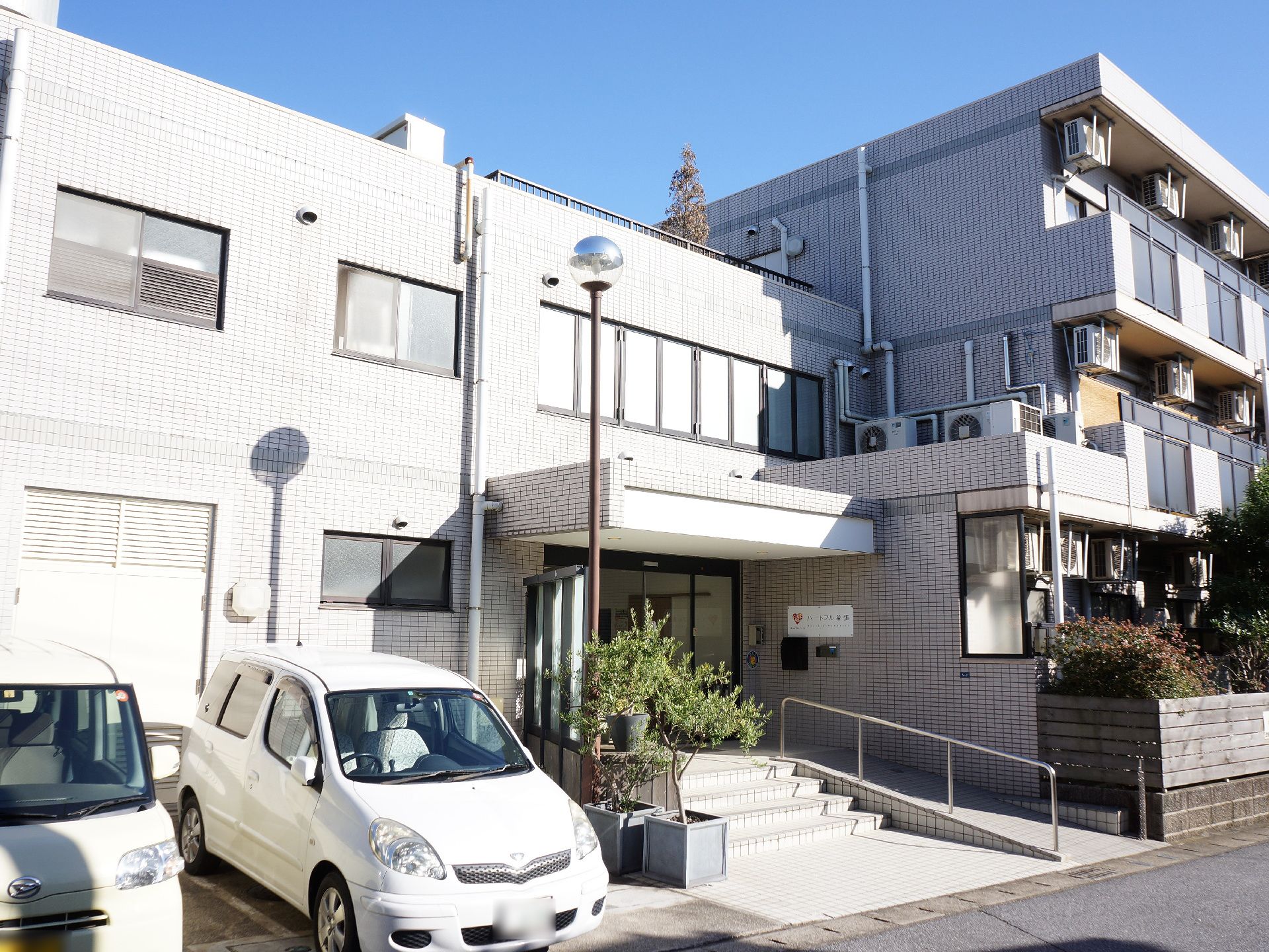 マンションのような外観となっている施設の外側は日の光を浴びており、スロープと階段のある入口の横側には数台の自動車がある。