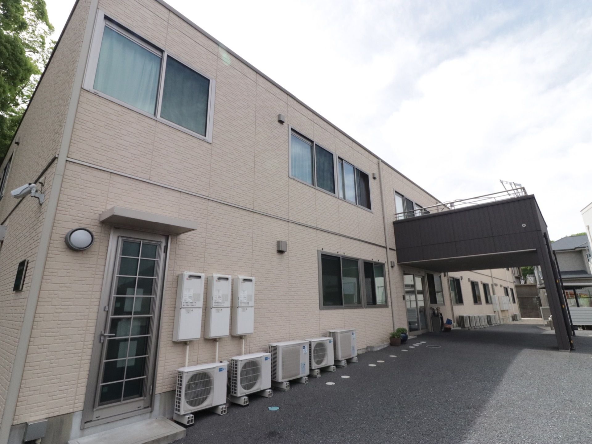 屋根付きの玄関の他にもドアが設置してある。給湯器や室外機が前面に置いてある。裏口方面には防犯カメラもある。