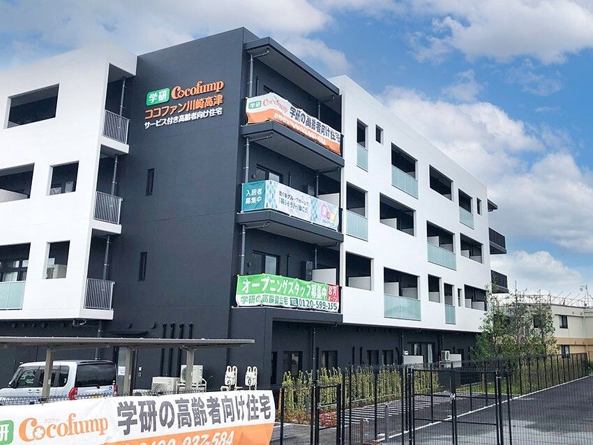 白とダークグレーの建物は4階建てで、敷地内には駐車場があり、建物の周りには草木が植えられている。