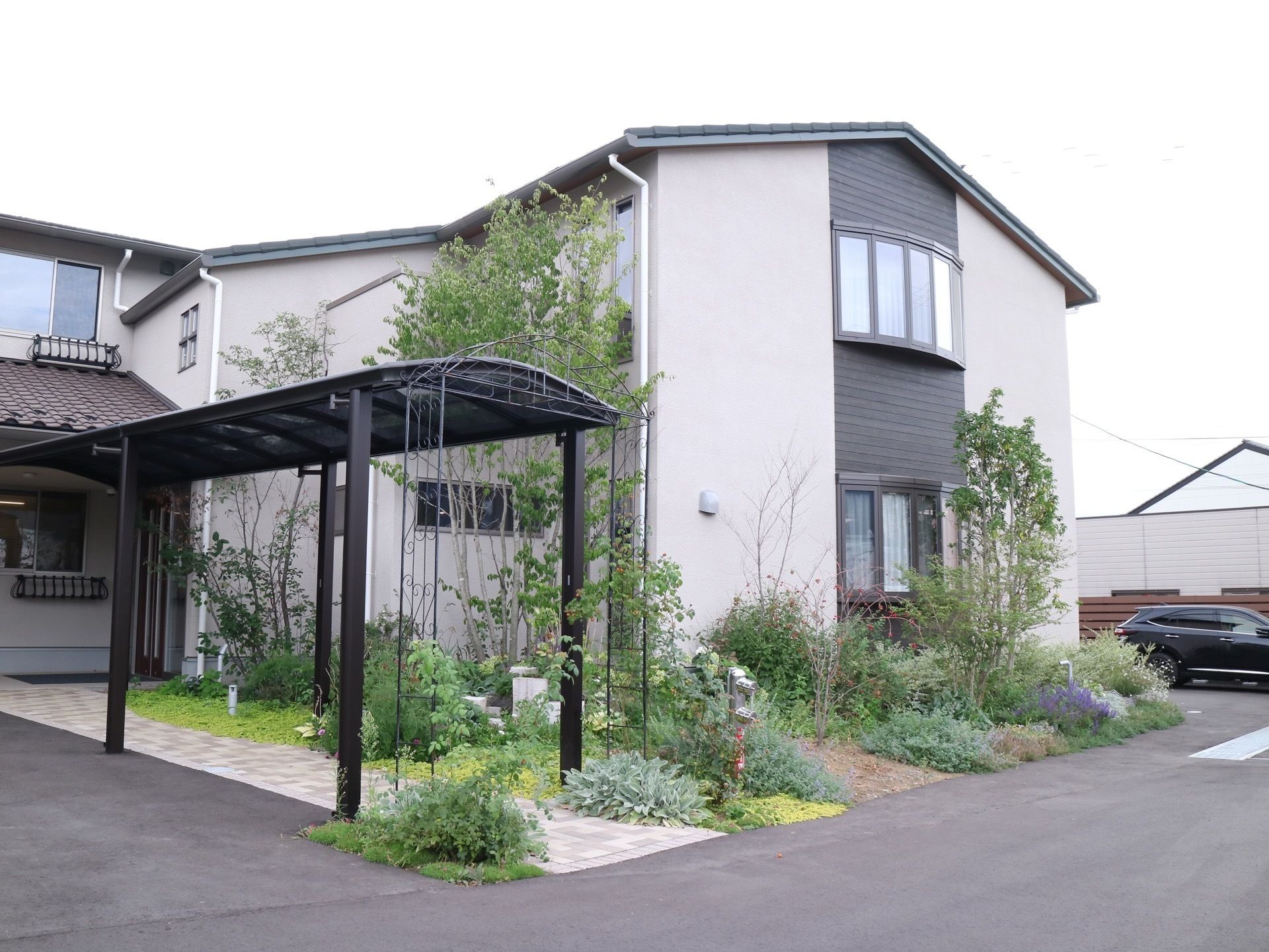 アスファルトで舗装された敷地に２階建ての住宅が建っている。玄関までは屋根の付いたアプローチがあり、植栽に囲まれている。