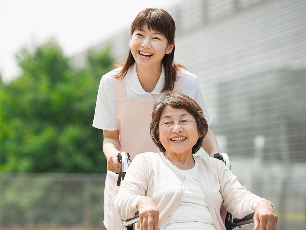 ピンクのカーディガンを着た車いすの高齢女性とピンクのエプロンを付けた若い女性スタッフが笑顔で散歩をしている。