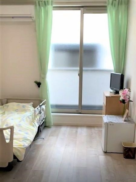 大きな窓があるフローリングの部屋には寝具付きのベッドや棚があり、花が飾られている。