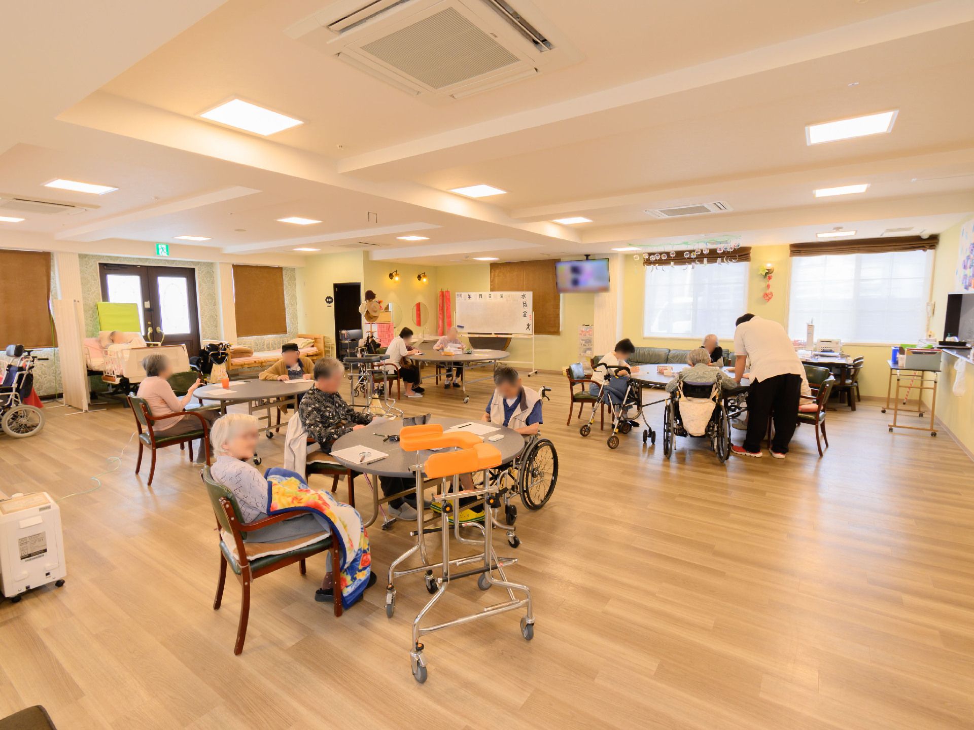 窓に囲まれた広間に配置されている丸テーブルの周りの椅子に座る入居者様