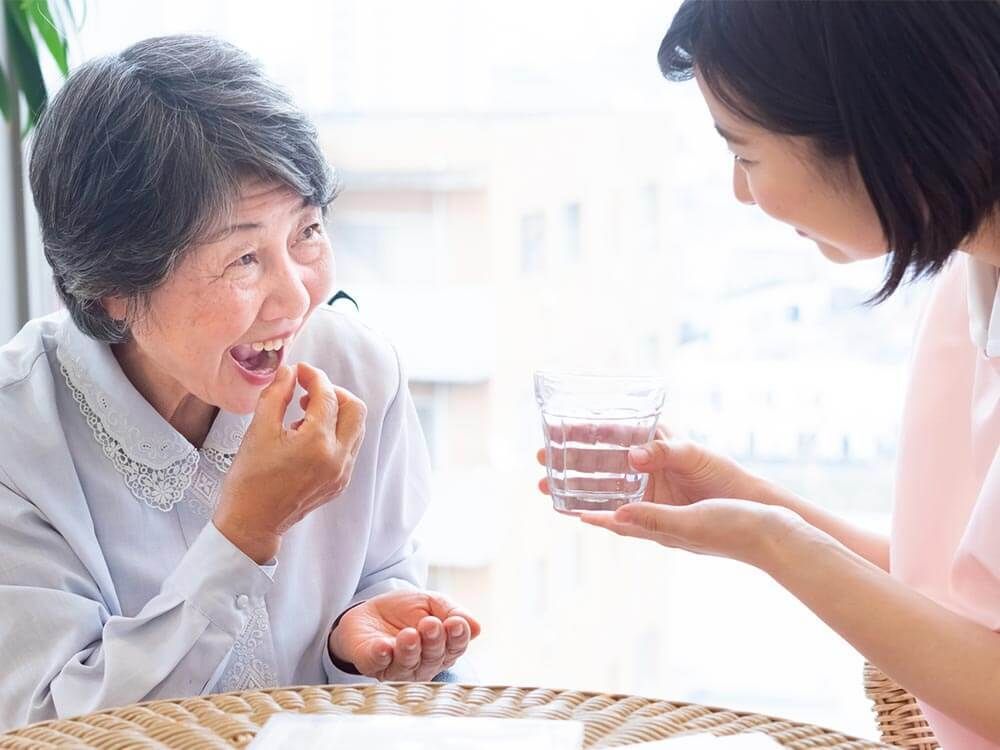 薬を飲もうとしている高齢女性と水の入ったコップを持っているエプロンを付けた女性スタッフ。