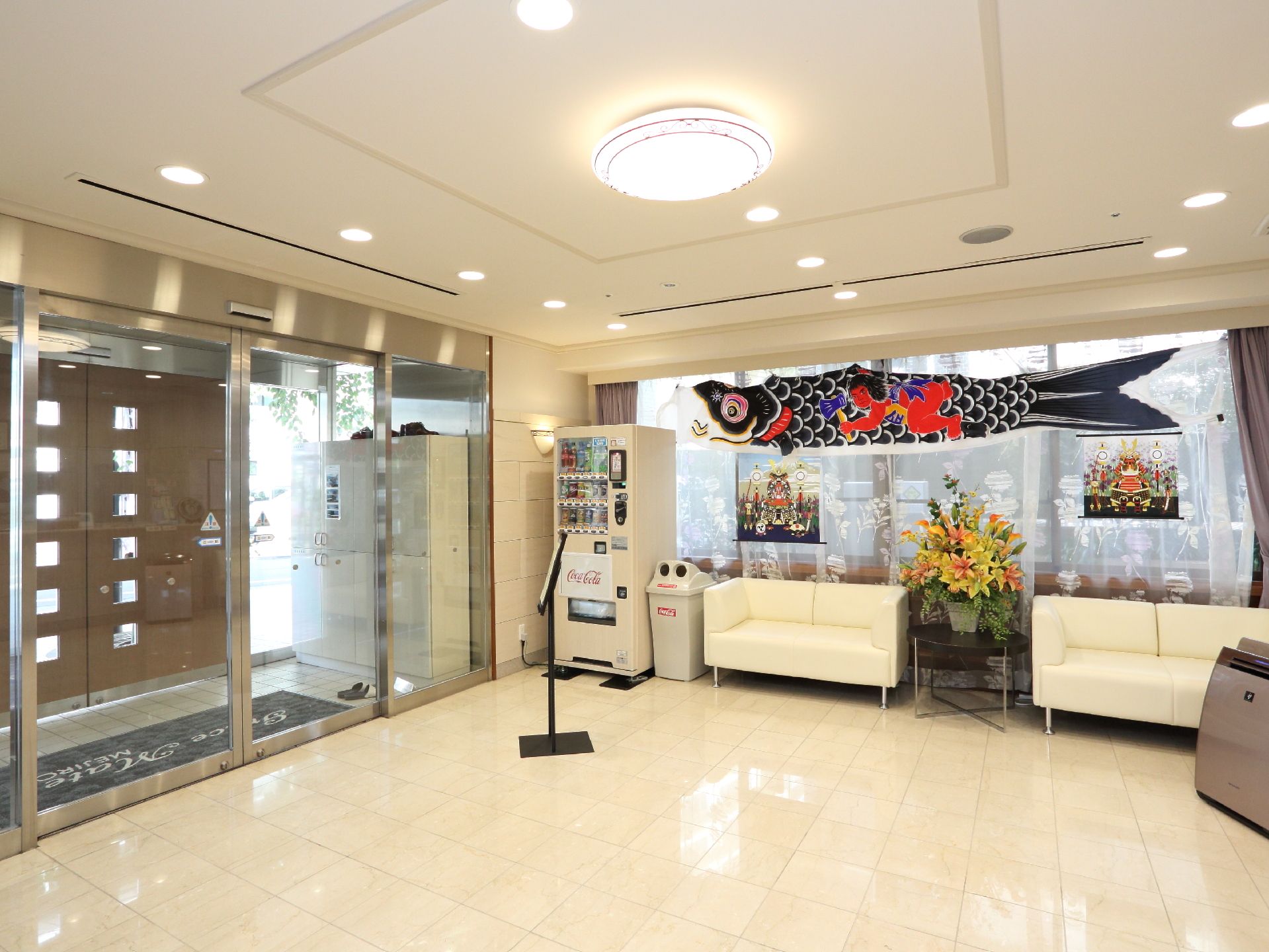 明るいエントランス。バリアフリー構造。脇にはソファー、大きな花、自動販売機が設置され、上部に鯉のぼりと端午の節句がテーマの絵がある。