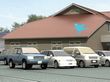 ブラウン系の建物が建っていて、屋根もブラウン。敷地内には広い駐車場があり、車が止まっている。