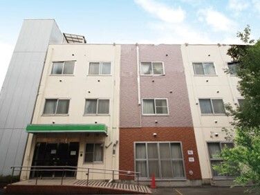 ベージュとブラウンの３階建ての建物。玄関先には手すり付きのスロープがあり、駐車場もある。