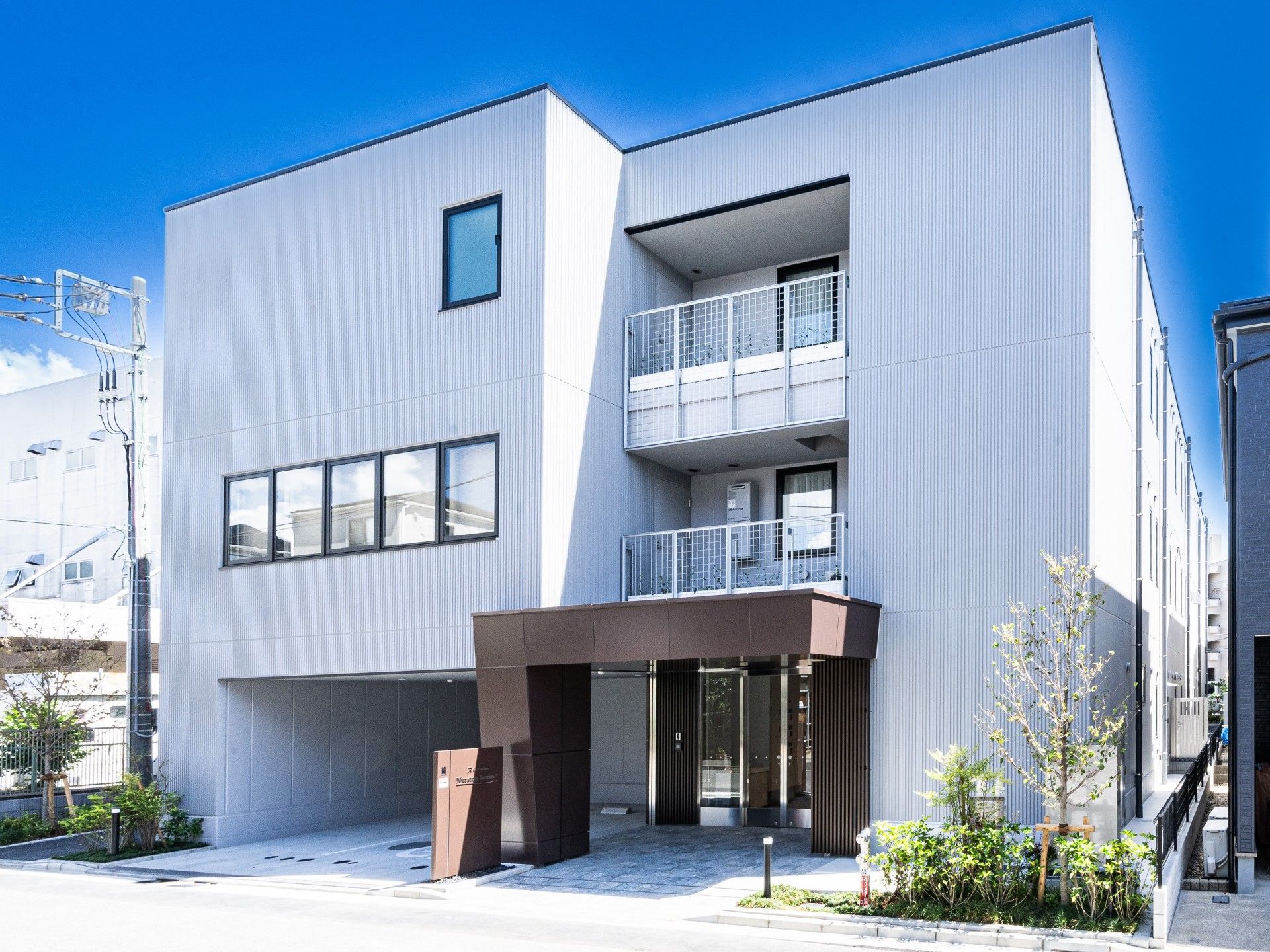 シルバー系の外壁の３階建ての建物は日当たりのいい場所に建っていて、各フロアにベランダがあり、草木も植えられている。