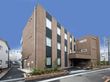 ブラウン系の外壁の3階建ての建物の玄関先には屋根があり、外壁にはホーム名が書かれている。
