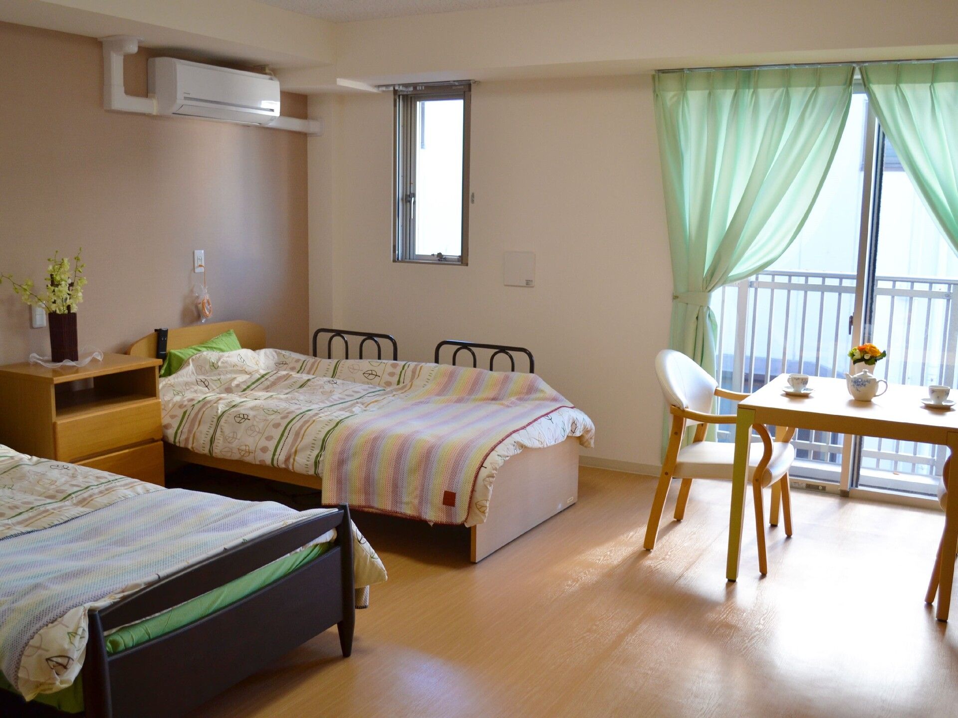 レースカーテンとカーテンが付いた大きな窓があるフローリングの部屋にはエアコンや寝具付きのベッドが2台ある。