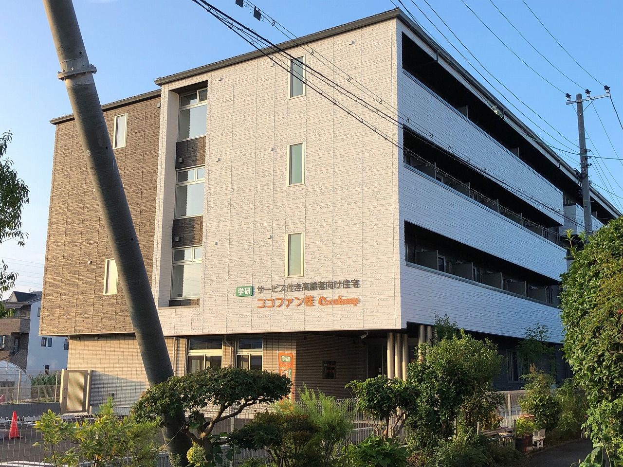 白とブラウンのエル字型の建物は4階建ての建物の建っている敷地内には草木も植えられている。
