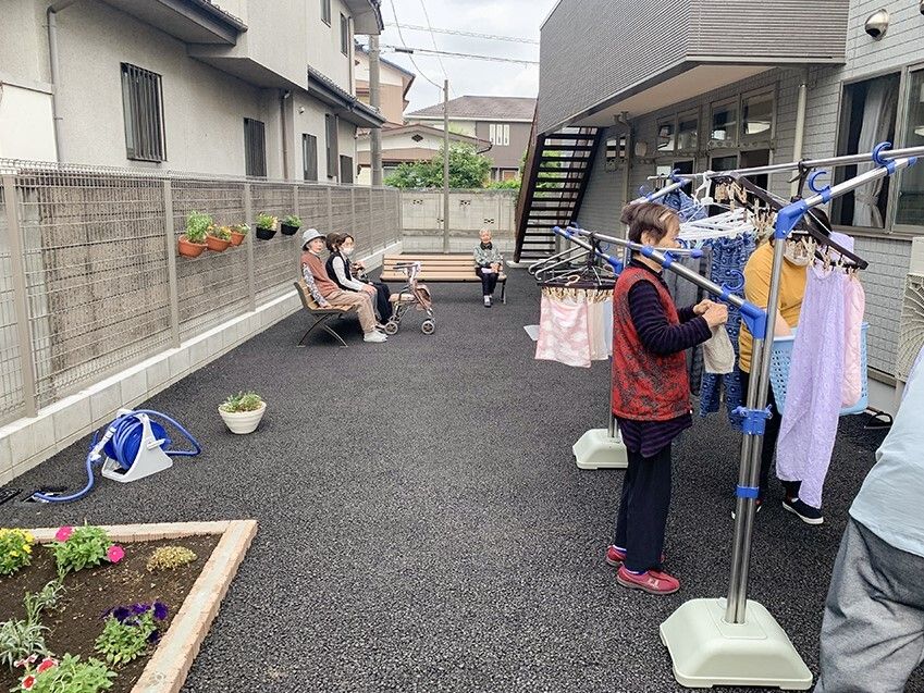 屋外のベンチに座って外気浴をしている高齢者たちと洗濯物を干している高齢者がいる。