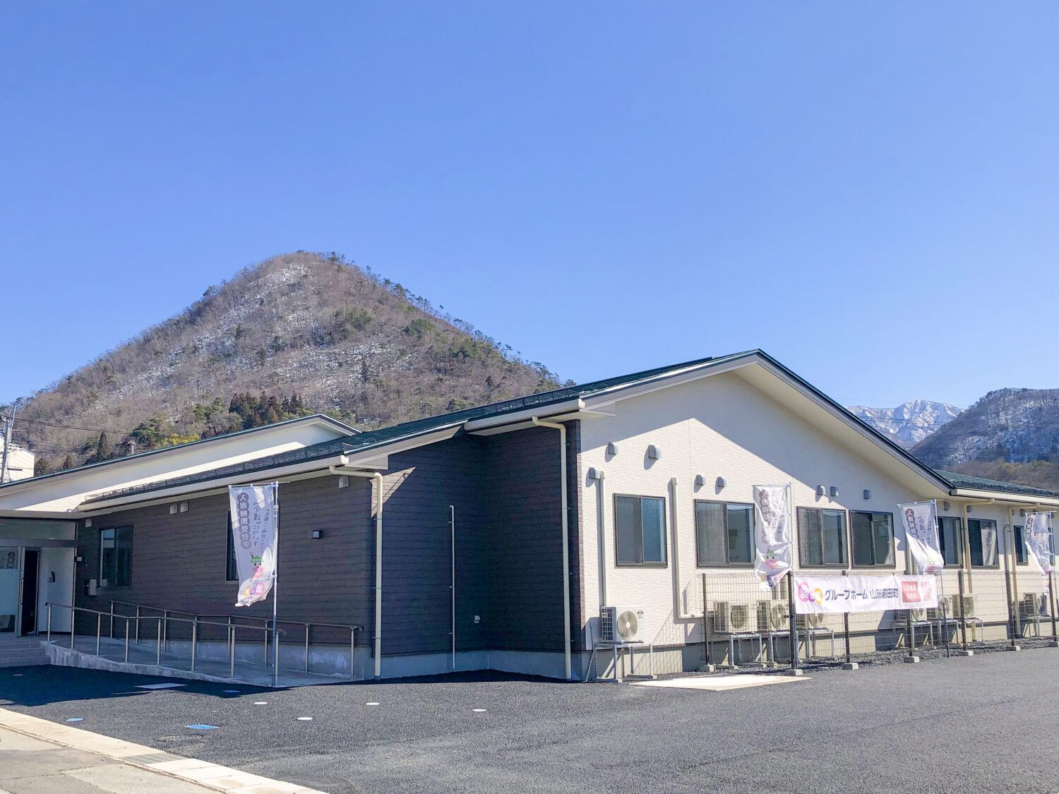 青い空の下に建っている平屋建ての建物の玄関には手すり付きのスロープがあり、建物の後ろには山がある。