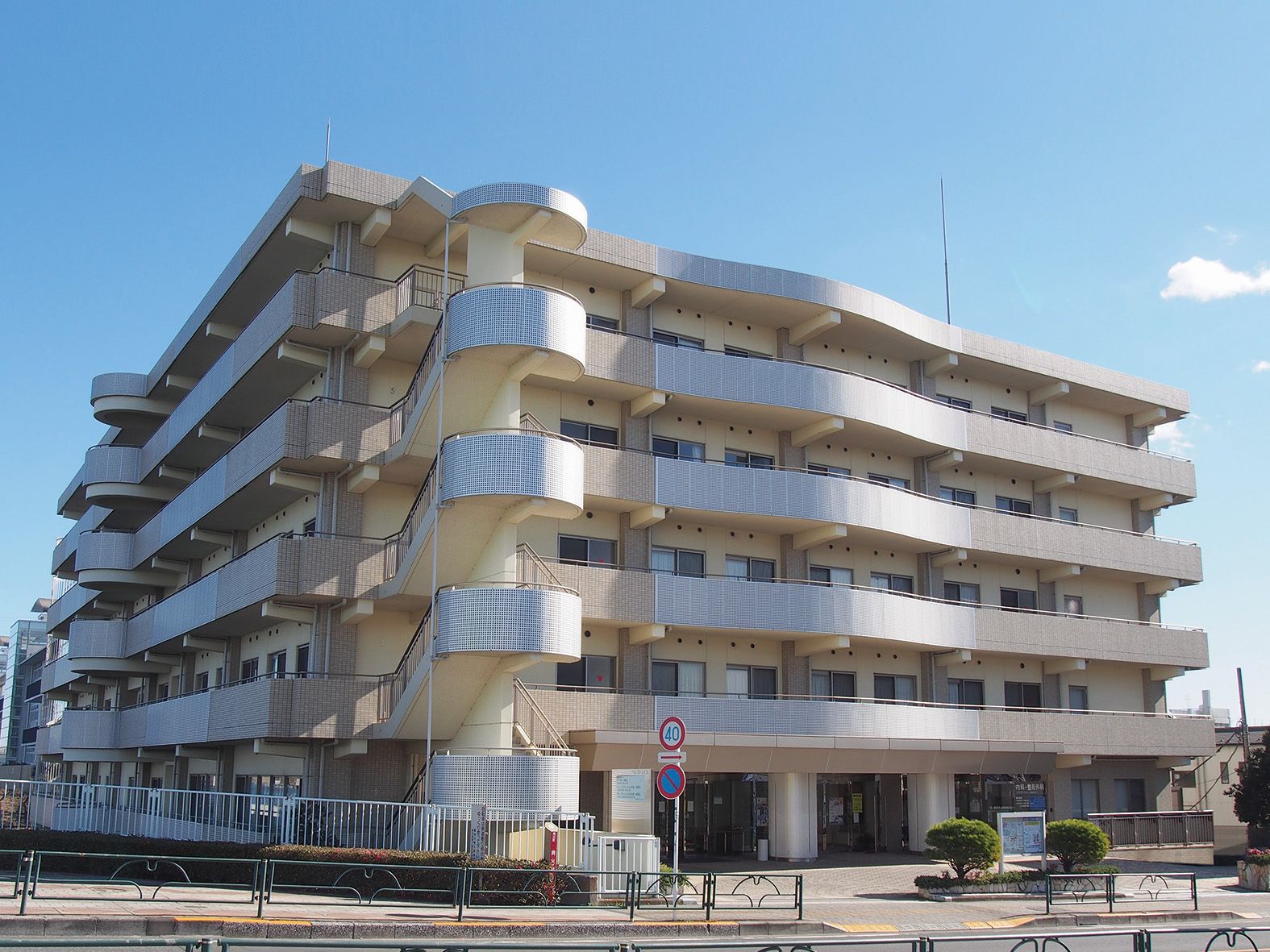 大きな道路に面した、5階建てのマンションのような外観である。周辺の道路には、ガードレールや横断歩道が設けられている。