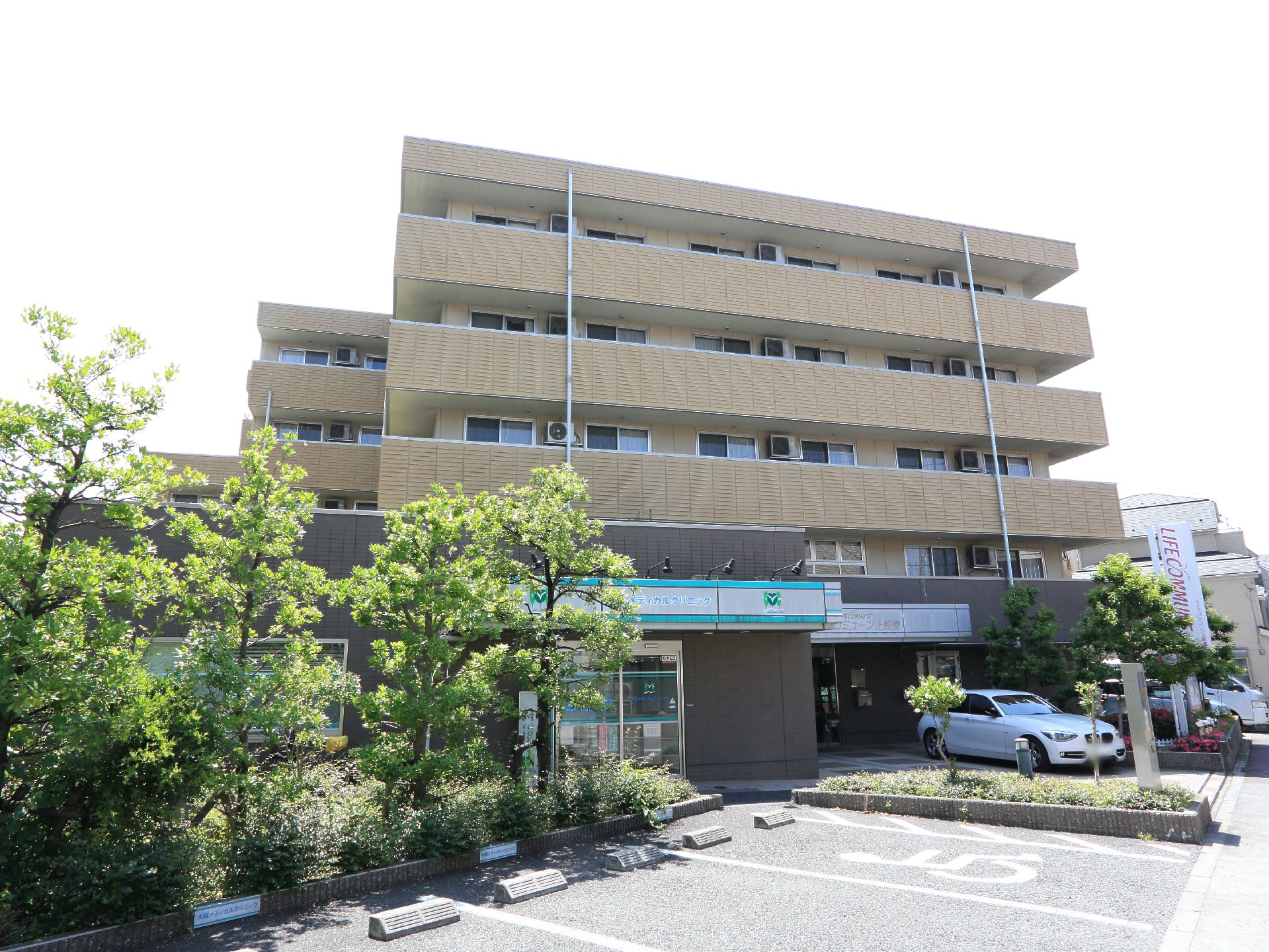 茶色の壁面の5階建の施設で目の前が駐車場になっている。コンビニが近くに立っており樹木も植えられている。