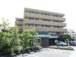 茶色の壁面の5階建の施設で目の前が駐車場になっている。コンビニが近くに立っており樹木も植えられている。