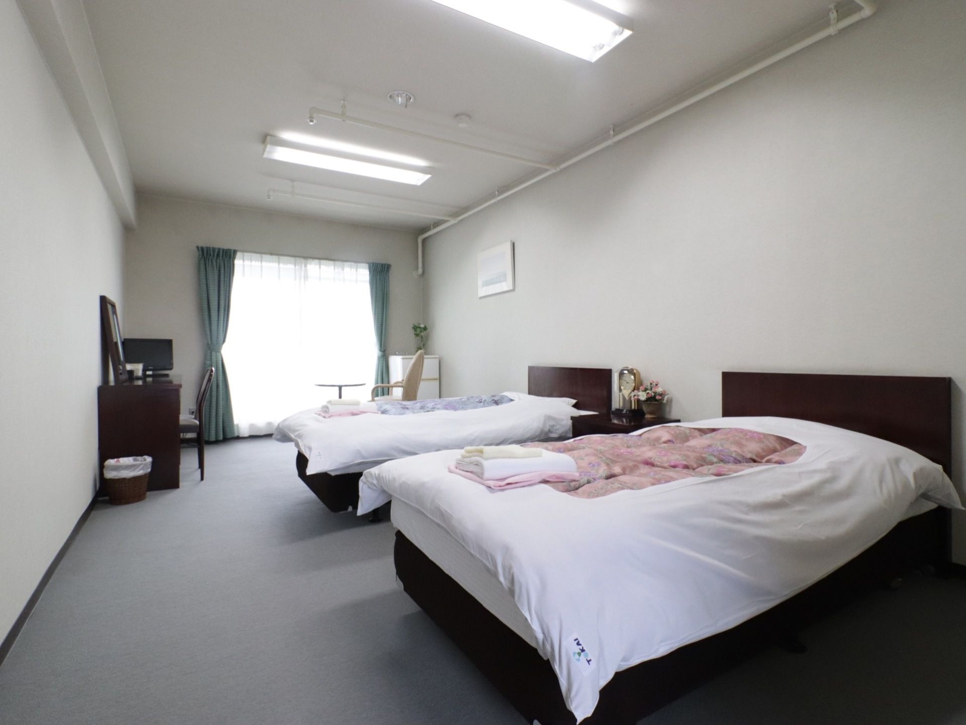 薄いグレーのフローリングを取り入れた一室にベッドを２台並べて配置している。正面のガラス戸際に冷蔵庫、テーブルセット、テレビなどを備えている。