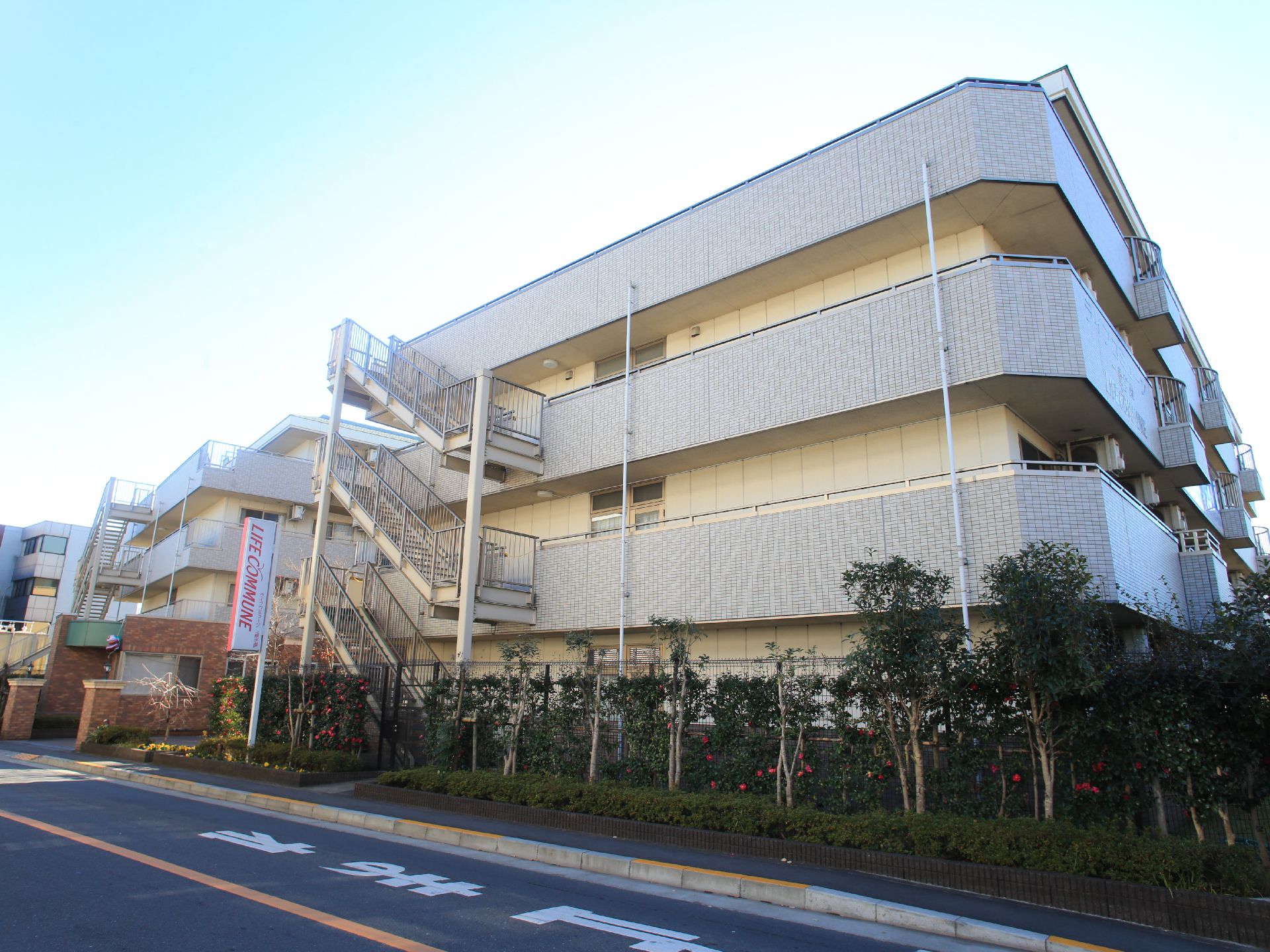 ベージュの落ち着いた雰囲気の建物は3階建てになっている。建物の周りの道路沿いには、木々が植えられている。